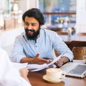 closeup-view-smiling-indian-business-man-caucasian-female-business-partners-sitting-together-table-examining-documents (1)